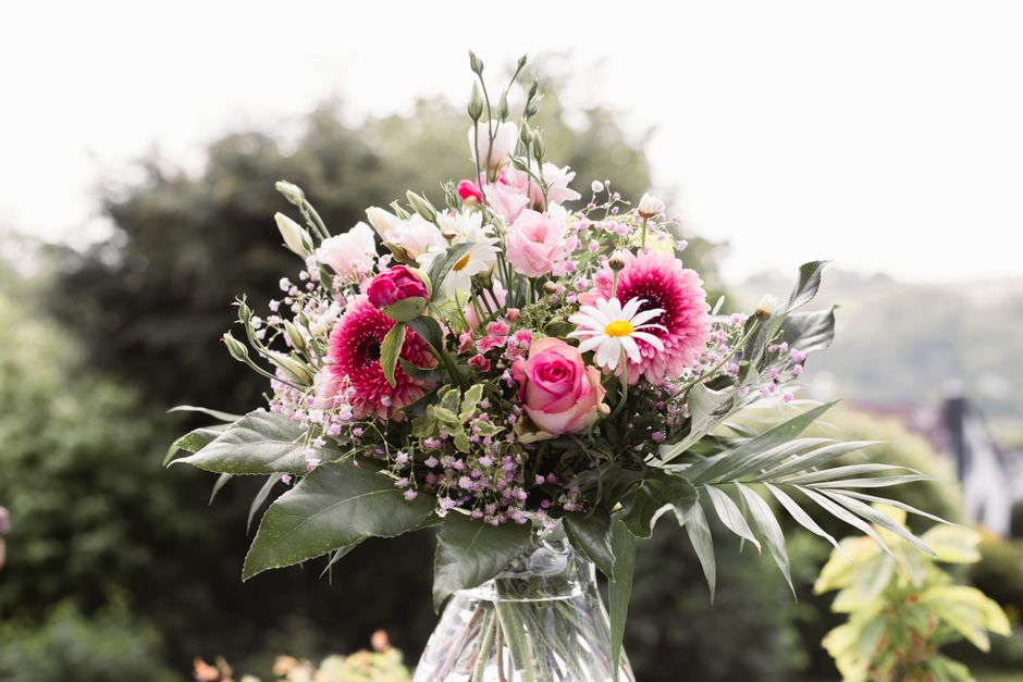 A vibrant floral bouquet featuring pink and white daisies, pink roses, and pink gerbera daisies arranged with lush green leaves and delicate filler flowers. The bouquet is displayed in a clear glass v